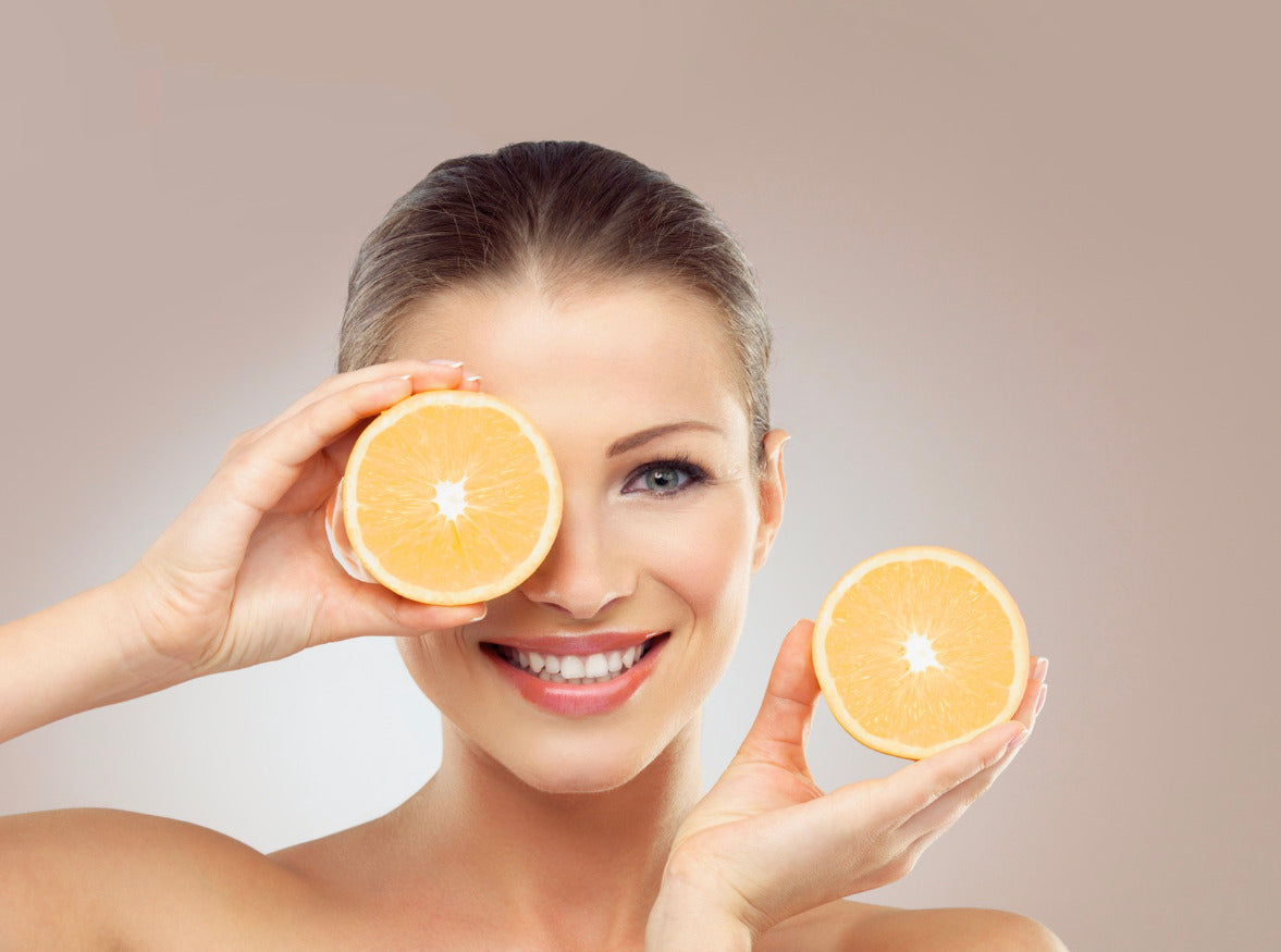 chica sonriente sosteniendo dos rodajas de naranja, una delante de su ojo y otra al costado de la cara, en alusion a la vitamina C